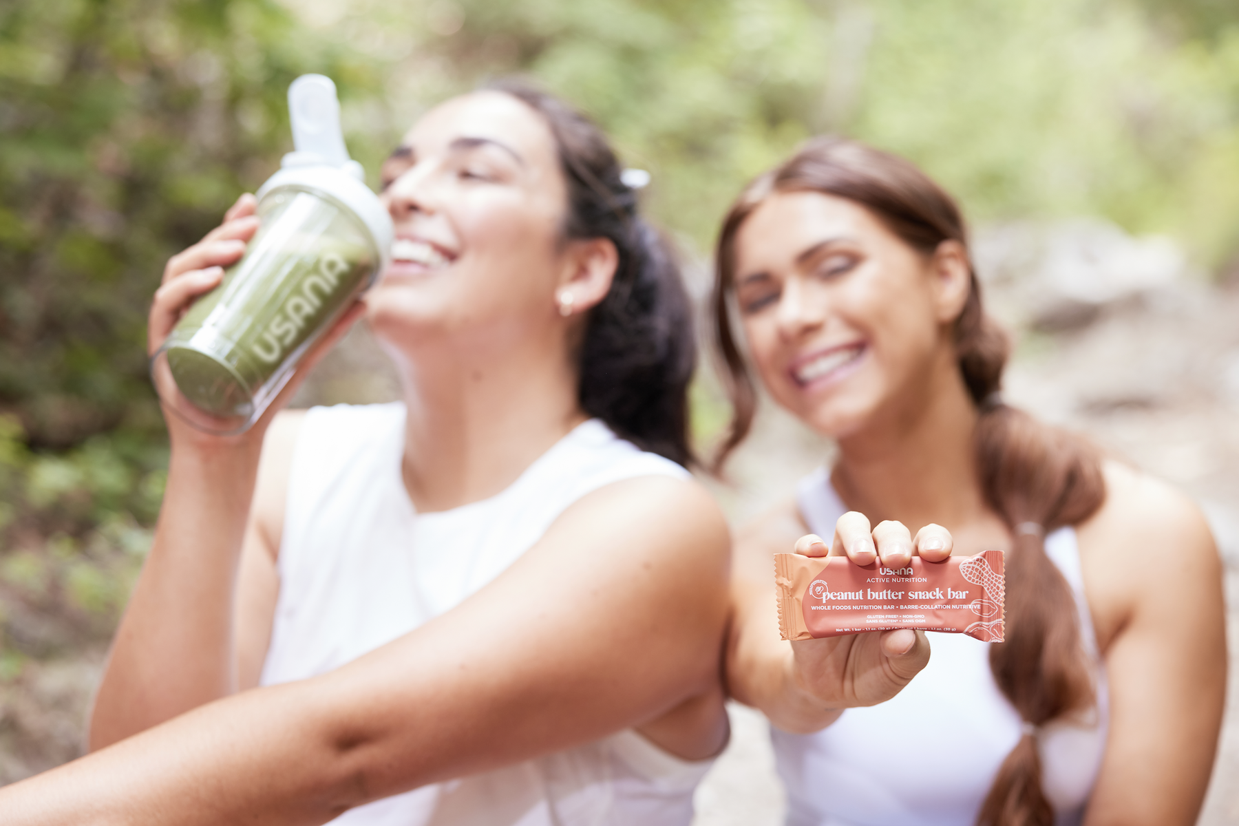 Two women smiling on hiking trail, one having a protein shake, the other showing a protein peanut butter snack bar.